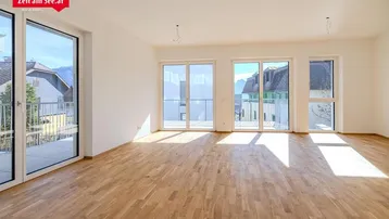 Heller, leerer Wohnraum mit Parkettboden, großen Fenstern und Zugang zum Balkon, mit Bergblick.