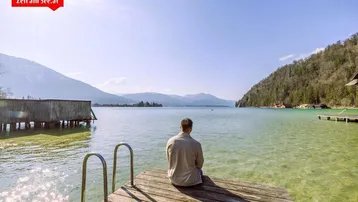 Person sitzt auf einem Holzsteg und genießt den weiten Blick über den See und die umliegenden Berge.