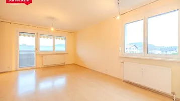 Helles, leeres Wohnzimmer mit Laminatboden, großen Fenstern und Zugang zum Balkon.