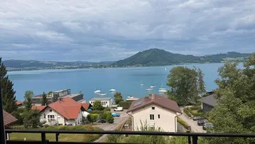 Blick von einem Balkon auf einen See mit Bergen und umliegenden Häusern unter bewölktem Himmel.