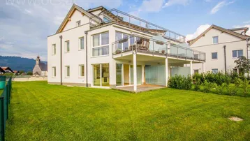 Modernes Wohngebäude mit großem Garten, Holzterrasse und Balkonen unter blauem Himmel.