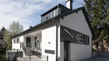 Modernes Mehrfamilienhaus mit weißer Fassade, dunklem Dach und Zugang zum Eingangsbereich.