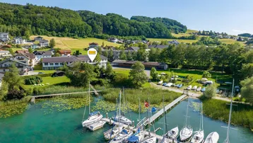 Luftaufnahme eines Anwesens am See mit Bootsanlegestelle, umgeben von grünen Hügeln und Wohnhäusern.