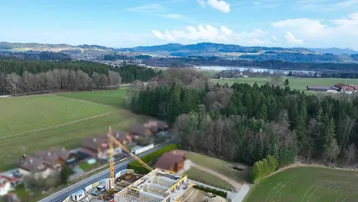 Luftaufnahme einer Baustelle mit Kran und Neubau in ländlicher Umgebung, umgeben von Feldern, Wäldern, einem See und Bergen unter blauem Himmel.
