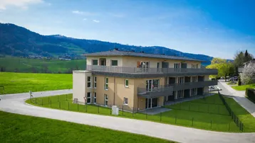 Modernes Mehrfamilienhaus mit Balkonen, umgeben von grünen Wiesen und Bergblick unter blauem Himmel.