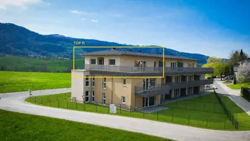 Modernes Mehrfamilienhaus mit Balkonen und Bergblick, umgeben von grüner Landschaft und Photovoltaikanlage.
