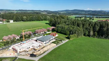 Luftaufnahme einer Baustelle mit Kran, umgeben von grünen Feldern und Wäldern mit Seeblick.