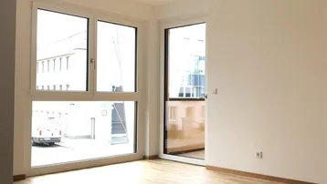 Heller, unmöblierter Wohnraum mit großen Fenstern, Holzboden und Zugang zu einem Balkon.