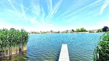 Idyllisches Seeufer mit kleinem Holzsteg und Schilf unter einem weiten blauen Himmel.