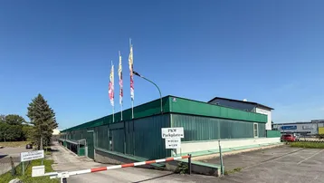 Außenansicht eines Gewerbegebäudes mit Parkplatz, Schranke und Fahnen unter blauem Himmel.