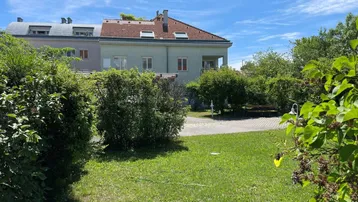 Blick auf die Gebäudefassade mit Gartenbereich, umgeben von üppigem Grün und einem blauen Himmel.
