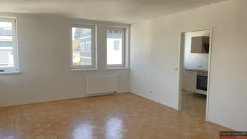 Helles Zimmer mit Parkettboden, weißen Wänden und Blick nach draußen durch zwei Fenster.