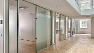 Heller Bürobereich mit Glaswänden, Holzparkett und Blick auf eine obere Etage.