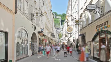 Belebte historische Gasse mit traditionellen Geschäften, vielen Passanten und Blick auf eine Bergfestung.