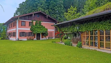 Traditionelles Landhaus mit rosafarbener Fassade, grüner Bepflanzung und überdachtem Essbereich im Freien.