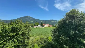 Weitläufiger Blick über grüne Felder und Bäume auf die umliegende Berglandschaft unter blauem Himmel.