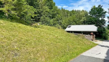 Kleines rustikales Gebäude am Hang, umgeben von Wald und Wiese, mit Holzstapel und Solaranlage.