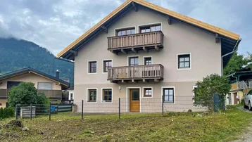 Traditionelles Haus mit Holzbalkonen und Bergblick in ländlicher Umgebung unter bewölktem Himmel.