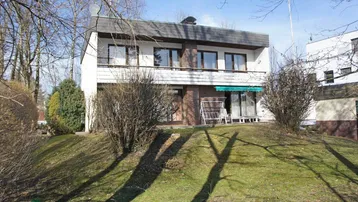 Freistehendes Haus mit weißer Fassade, Balkon und Gartenbereich auf einem sonnigen Grundstück.
