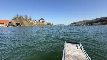 Weitläufiger Blick über den See mit umliegender Landschaft und Gebäuden am Ufer unter blauem Himmel.