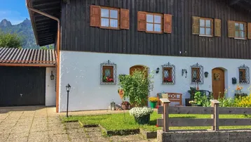 Traditionelles Haus mit Holzelementen und weißer Fassade, umgeben von einem kleinen Vorgarten und Bergblick.
