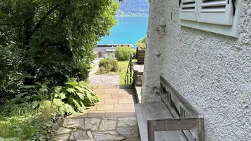 Steinweg führt zu einem Seeufer mit einer Holzbank neben dem Haus, umgeben von üppigem Grün.