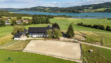 Luftaufnahme eines Anwesens mit Reitplatz, Stallungen und malerischem Seeblick in ländlicher Umgebung.