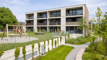 Modernes Mehrfamilienhaus mit Balkonen, umgeben von einer gepflegten Grünanlage und einem Kinderspielplatz.