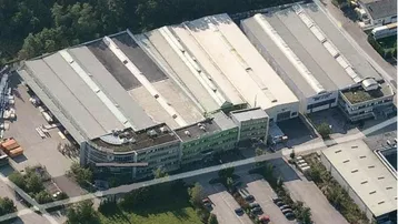 Luftaufnahme eines großen Gewerbekomplexes mit mehreren Hallen, Bürogebäuden und Parkplätzen, umgeben von Bäumen und Grünflächen.