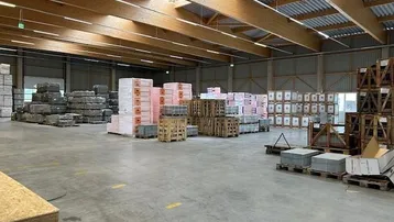 Geräumige Lagerhalle mit hohen Holzdecken, Oberlichtern und gestapelten Baumaterialien auf Paletten.