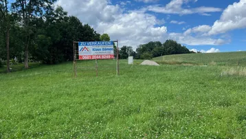 Verkaufsschild auf einem grünen Grundstück unter bewölktem Himmel mit Bäumen im Hintergrund.