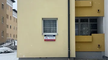 Gelbe Fassade eines Mehrfamilienhauses im Winter mit einem 'Zu Kaufen'-Schild am Fenster.