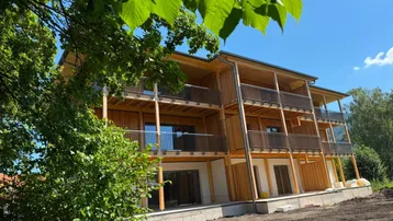 Neubau mit Holzfassade und mehreren Balkonen, umgeben von viel Grün unter blauem Himmel.