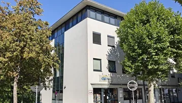 Modernes Bürogebäude mit weißer Fassade und großen Fenstern, umgeben von Bäumen an einer Straße.