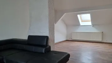 Geräumiges Wohnzimmer mit schwarzem Sofa, Holzboden und Dachfenster, das viel Licht hereinlässt.