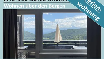 Blick aus dem Fenster eines Wohnraums auf einen Balkon und die umliegende Berglandschaft.