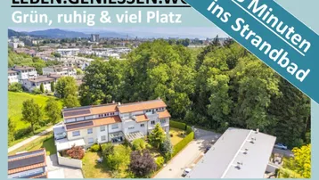Luftaufnahme eines Mehrfamilienhauses mit Garten, umgeben von viel Grün und Bergen im Hintergrund.