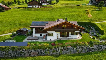 Luftaufnahme eines traditionellen Hauses mit Solaranlagen, umgeben von einem gepflegten Garten und grünen Hügeln.