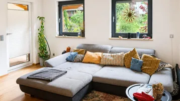 Gemütliches Wohnzimmer mit großem Ecksofa, Holzboden und Blick ins Grüne durch die Fenster.