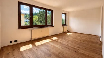 Helles Zimmer mit Holzboden und großen Fenstern, die viel Tageslicht hereinlassen und einen grünen Ausblick bieten.