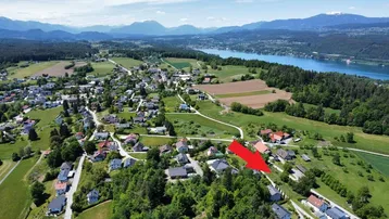 Luftaufnahme eines Dorfes mit See- und Bergblick, das Grundstück ist mit einem roten Pfeil markiert.