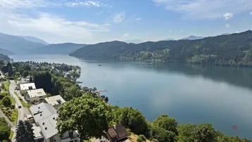 Panoramablick auf einen malerischen See, umgeben von grünen Hügeln und modernen Wohnhäusern am Ufer.