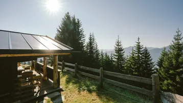 Sonnige Terrasse mit Holztisch und Bänken, umgeben von alpiner Natur und mit Solaranlage.
