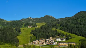 Panoramablick auf ein Bergresort mit mehreren Gebäuden und grünen Skipisten in alpiner Landschaft.