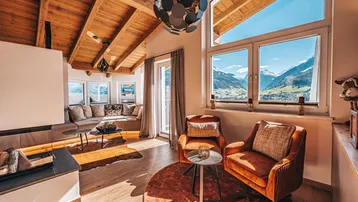 Helles Wohnzimmer im alpinen Stil mit Holzbalkendecke, Kamin und Bergblick durch große Fenster.