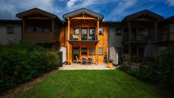 Holzverkleidetes Reihenhaus mit Terrasse und Garten, umgeben von grüner Landschaft.