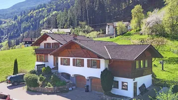 Traditionelles Haus im alpinen Stil mit Holzbalkonen und Satteldach, eingebettet in eine grüne Berglandschaft.