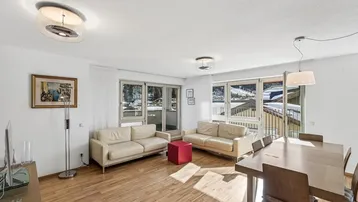 Helles Wohn- und Esszimmer mit Holzboden, beigen Sofas und Zugang zum Balkon mit Bergblick.