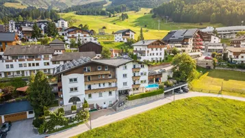 Luftaufnahme eines Hotels im alpinen Stil, umgeben von grünen Hügeln und Bergen.