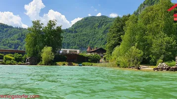 Malerisches Seeufer mit grünen Bergen im Hintergrund und Gebäuden am Ufer unter blauem Himmel.